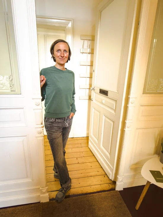Therapeutin steht entspannt an der Eingangstür der Praxis, eine Hand in der Hosentasche. Sie schaut leicht seitlich, die Altbauarchitektur mit verzierten weißen Türen ist im Hintergrund sichtbar.