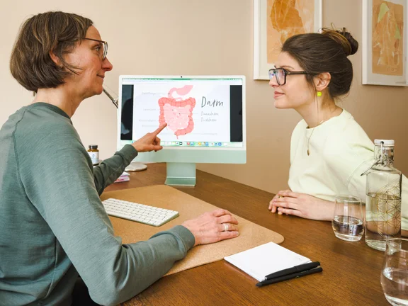 Therapeutin zeigt der Patientin am Schreibtisch auf einem grünen iMac eine anatomische Illustration des Darms mit deutschen Beschriftungen wie „Darm", „Dünndarm" und „Dickdarm". Die Patientin schaut aufmerksam auf den Bildschirm.