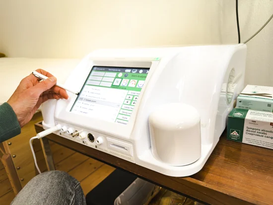 Nahaufnahme eines weißen medizinischen Analysegeräts (RAH-Analyse) auf einem Holztisch. Auf dem Touchscreen sind Menüpunkte zu sehen. Links hält eine Hand einen Eingabestift. Daneben liegen Alkoholtupfer in einer grünen Verpackung.