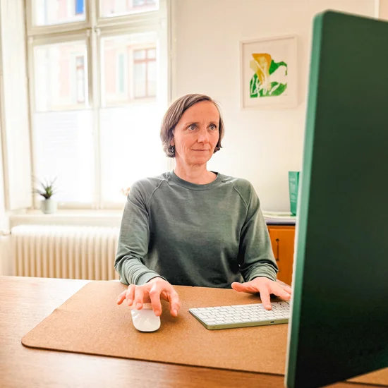 Therapeutin sitzt am Schreibtisch und bedient gleichzeitig Maus und Tastatur. Sie schaut konzentriert seitlich. Links im Bild ist das Behandlungsgerät sichtbar, hinter ihr ein Altbaufenster mit Pflanze und Blumenstrauß sowie ein Gemälde.