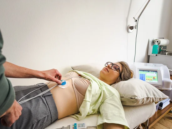 Nahaufnahme der liegenden Patientin mit einer einzelnen blauen Elektrode am Bauch. Die Therapeutin justiert mit einer Hand den Kabelanschluss. Rechts im Hintergrund ein medizinisches Analysegerät mit Touchscreen-Display.