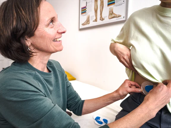 Therapeutin befestigt Elektroden-Pads auf dem Oberkörper der liegenden Patientin, die die Augen geschlossen hat. Mehrere blaue Elektroden sind bereits angebracht und mit Kabeln verbunden. Im Hintergrund ein anatomisches Poster an der Wand.