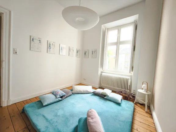 Ruhiger Yogaraum mit weißen Wänden und warmem Holzdielenboden. Im Mittelpunkt liegt eine türkisfarbene Yogamatte mit mehreren Entspannungskissen in Grau, Weiß und Blau für eine Restorative- oder Yin-Yoga-Einheit. An der Wand hängen anatomische Schautafeln des menschlichen Skeletts. Ein großes Sprossenfenster lässt natürliches Tageslicht herein. Neben dem Heizkörper steht ein kleiner weißer Beistelltisch mit einer Lampe. Die Atmosphäre wirkt ruhig und einladend.